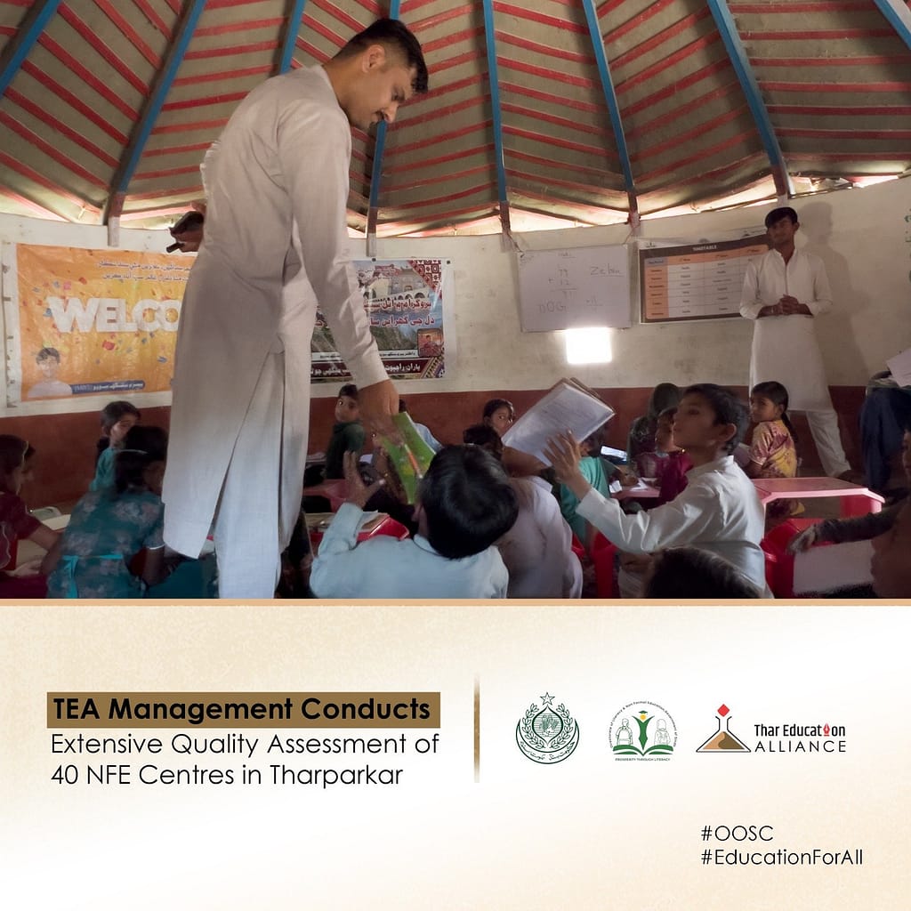 A man in white traditional attire stands on a chair, handing out books to students in a classroom with posters on the wall and a striped roof above.